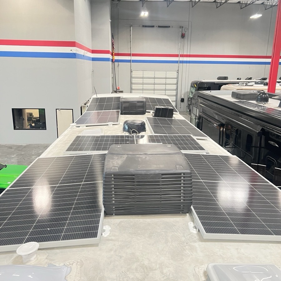 Solar panels mounted across the roof of a motorhome inside the 24/7 MotorCoach shop, with rooftop air conditioners and vents set between the panels and another coach parked alongside.